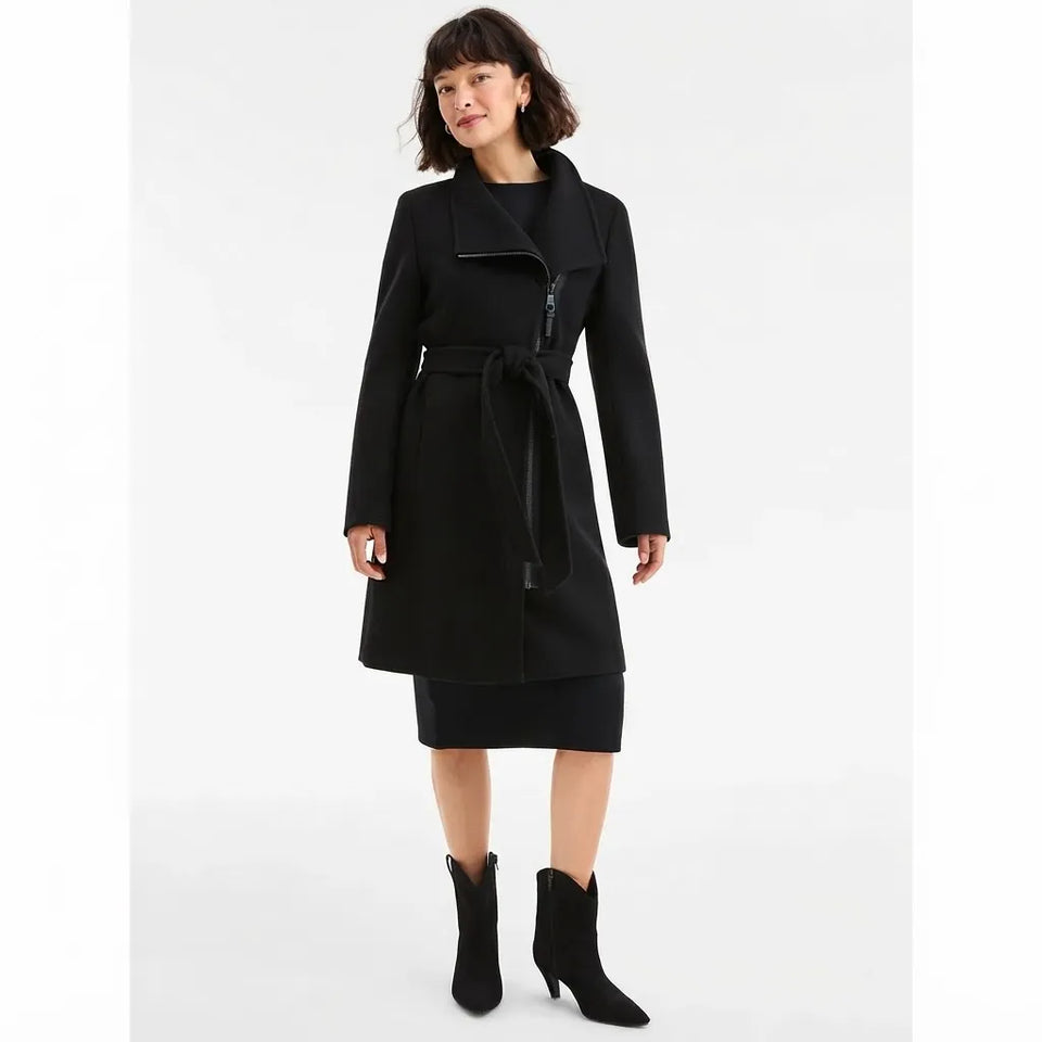 A woman modeling a black wrap-style coat with a zip front, tie belt, and notched lapels, paired with a black skirt and black ankle boots, against a plain light gray studio background.