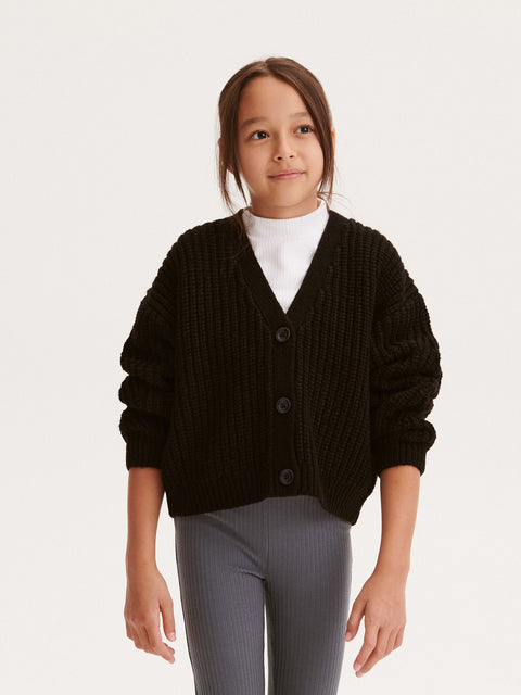 Child wearing a black cardigan over a white shirt with gray pants on a light gray background