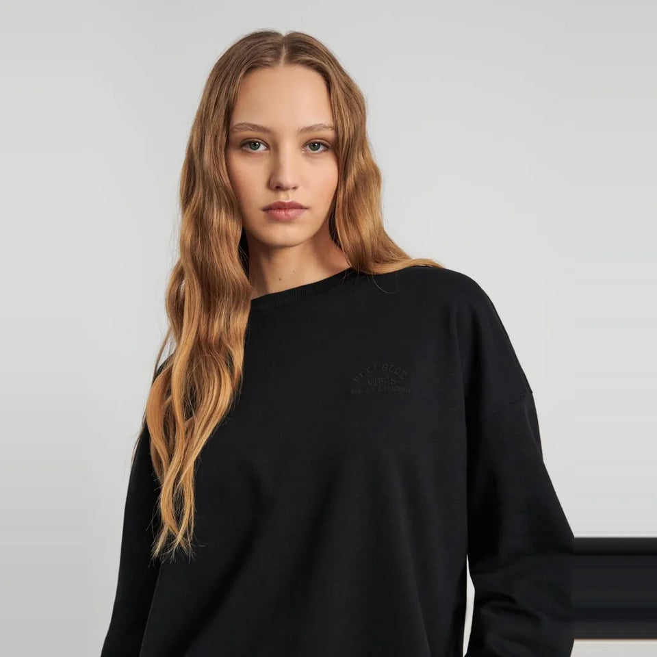 A woman with long blonde hair wearing a black crewneck sweatshirt, photographed against a light gray studio background.