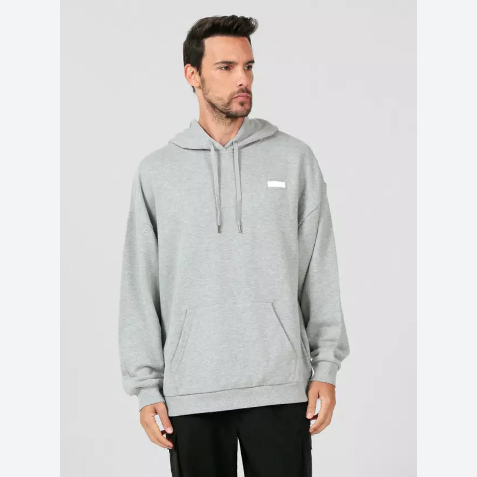 A man wearing a light gray heather pullover hoodie with a small white rectangular patch on the left chest, paired with black pants, set against a plain light gray studio background.