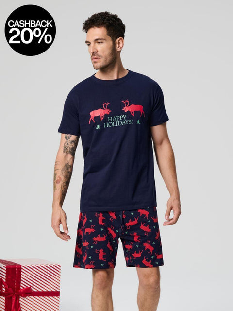 Man wearing navy t-shirt with reindeer design and matching shorts, standing next to a gift box on a light gray background.