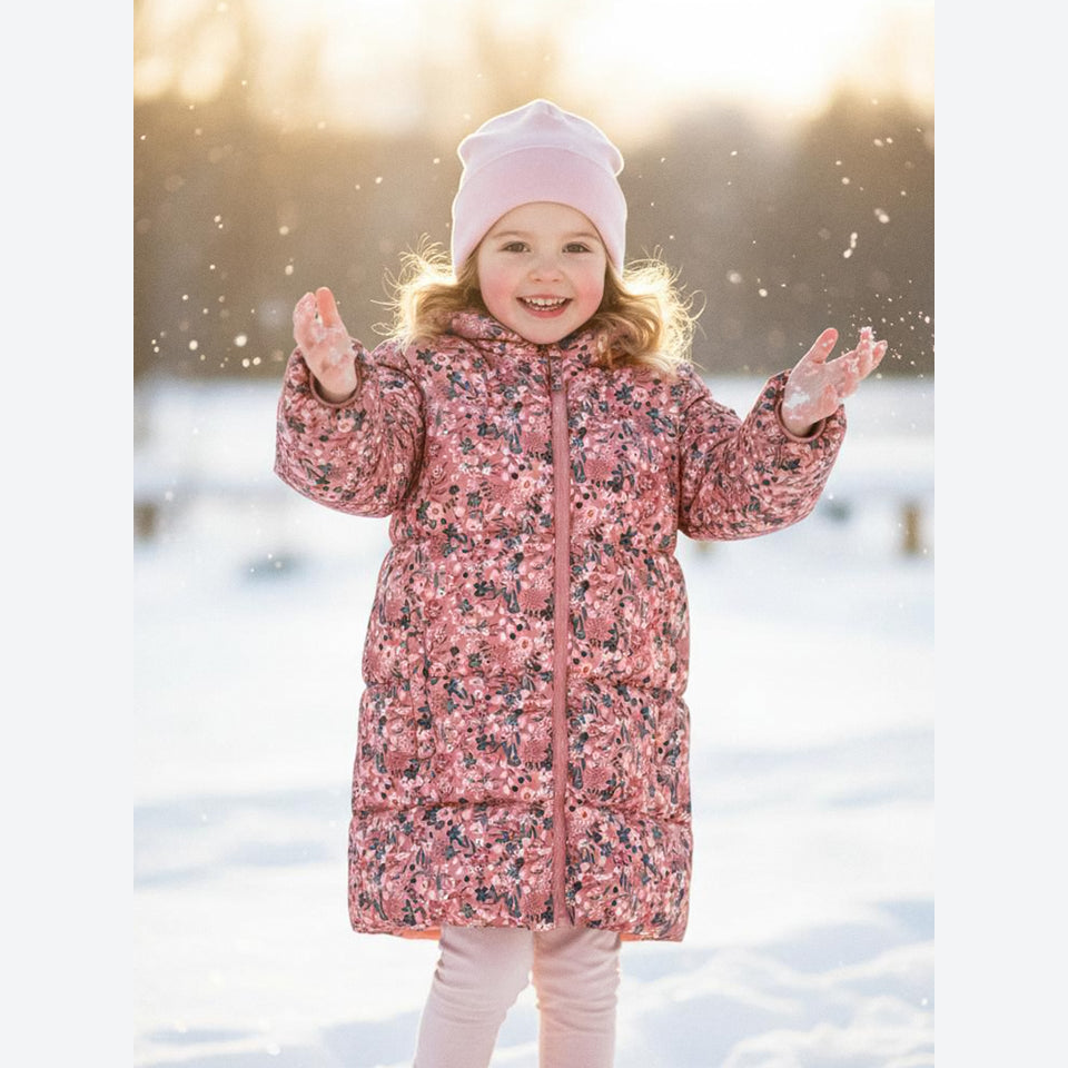 A young girl smiling in a snowy outdoor setting, wearing a pink floral puffer coat and a light pink beanie.