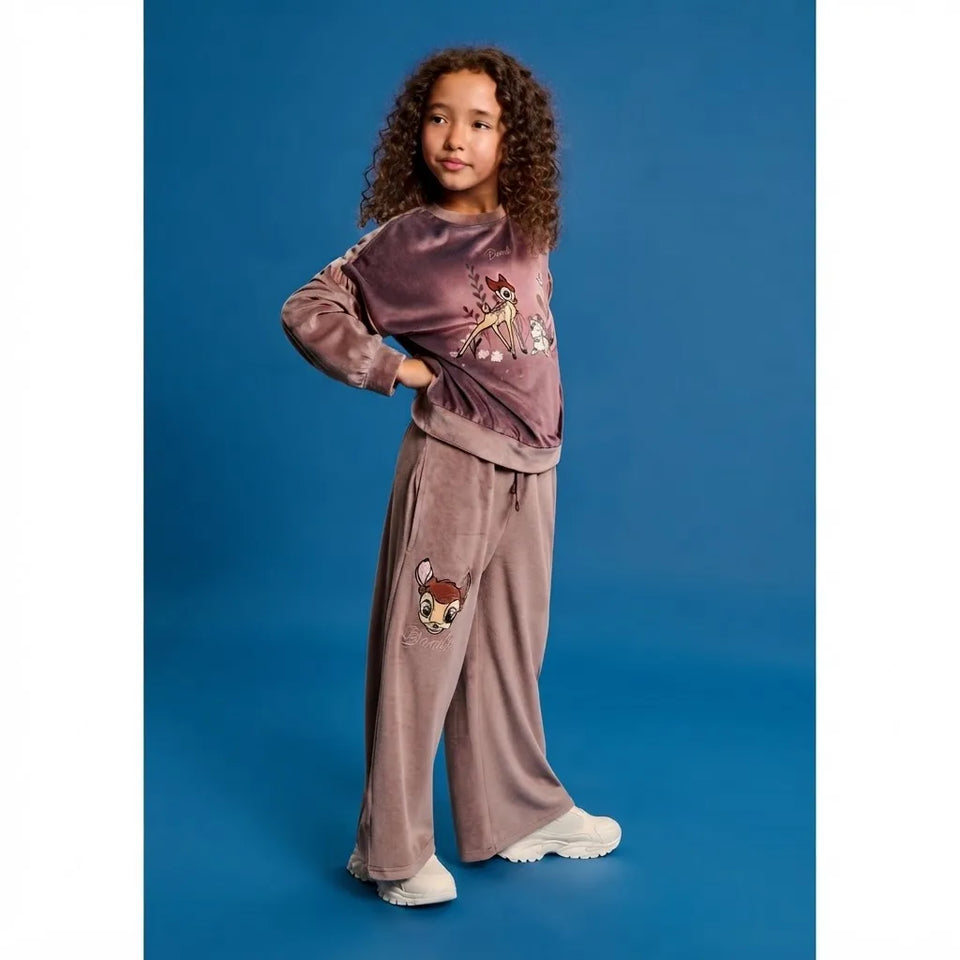 A young girl with curly brown hair stands against a solid blue studio background wearing a matching two-piece pajama set. The set consists of a long-sleeve top and wide-leg pants in a dusty mauve-purple velvet fabric, both decorated with cartoon animal graphics.