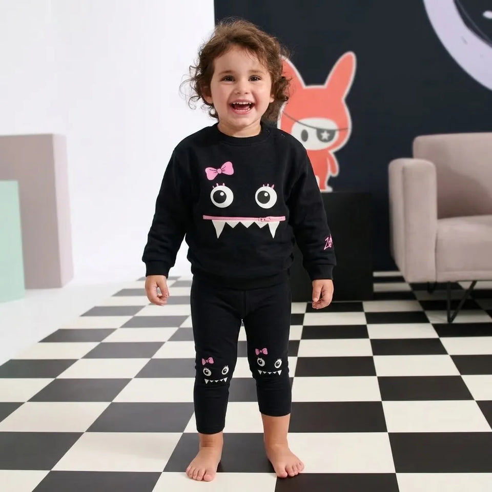 A toddler stands barefoot on a black-and-white checkered floor wearing a black two-piece pajama set with a monster face graphic on the sweatshirt and matching pants.