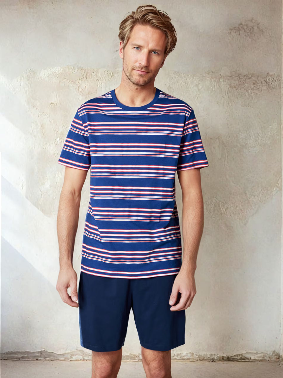 Man wearing a blue and red striped t-shirt and navy shorts against a textured wall.