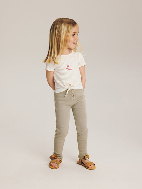 Young girl wearing a white t-shirt and beige pants on a plain background