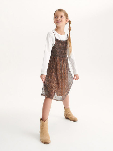 Young girl wearing a brown patterned dress over a white shirt on a white background