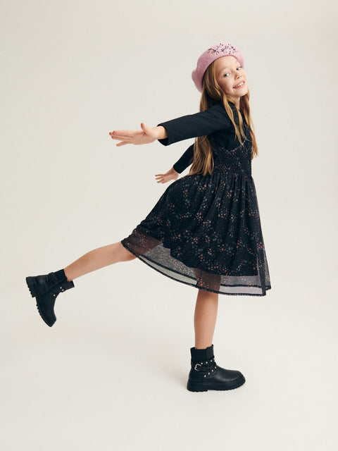 Young girl in a black dress with floral pattern and pink hat, posing against a white background