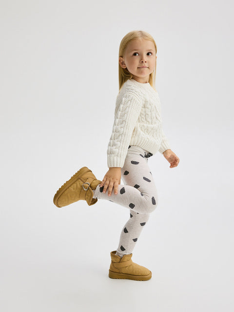 Child wearing a white cable knit sweater, polka dot leggings, and tan boots on a white background
