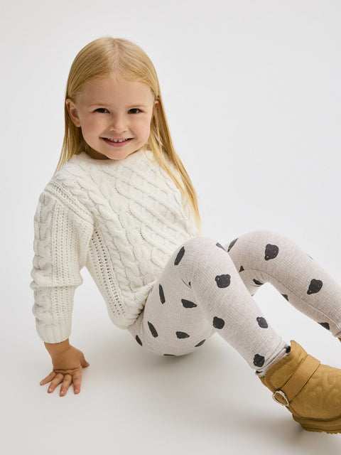 Child wearing a white cable knit sweater and polka dot leggings on a white background