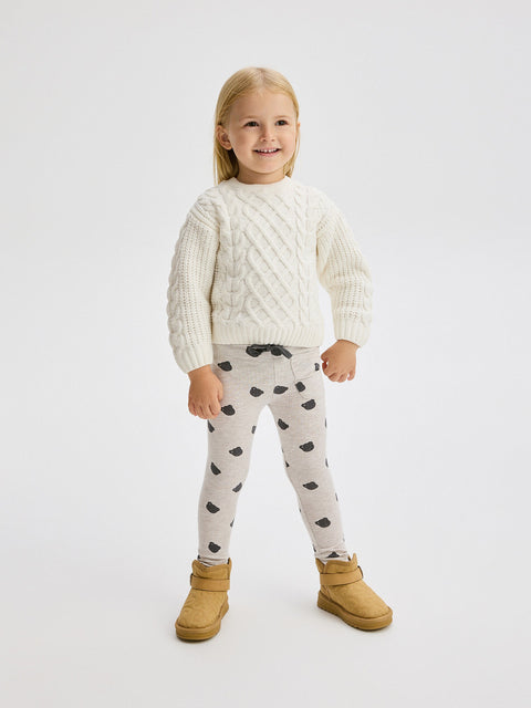 Child wearing a white cable knit sweater and patterned leggings on a white background