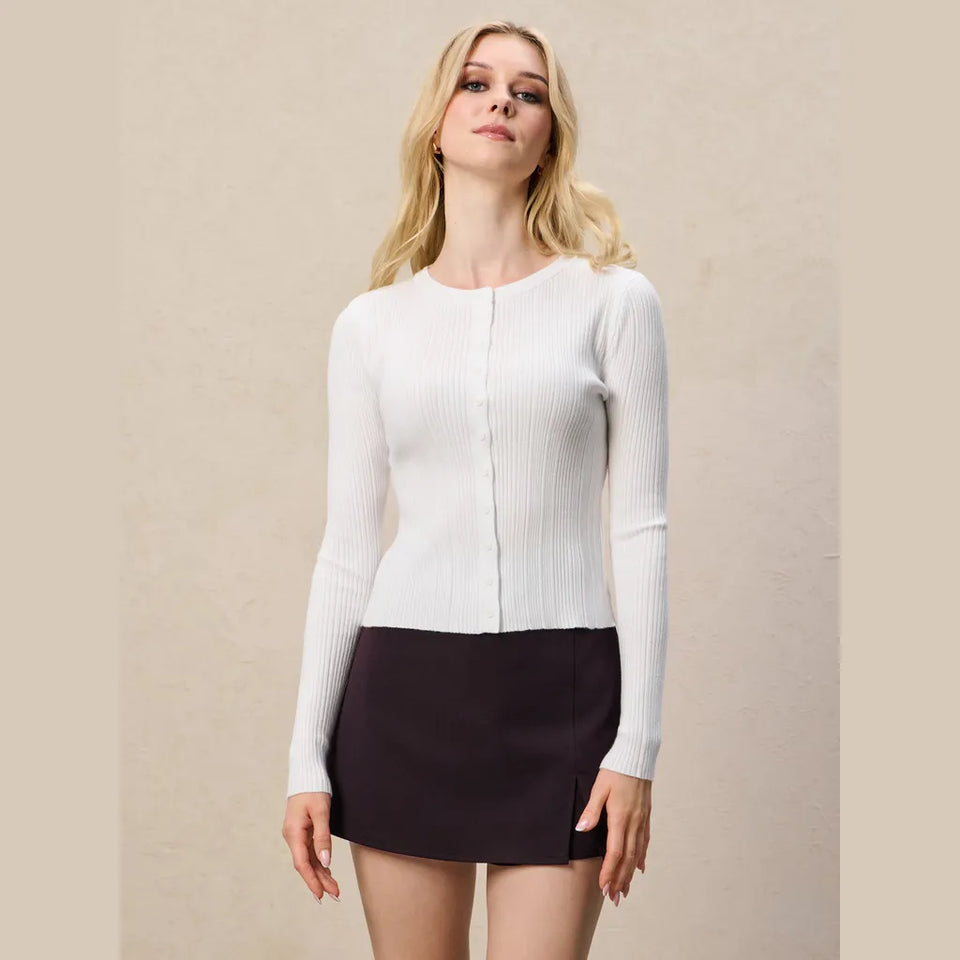 A blonde woman modeling a fitted white ribbed long-sleeve top with a front button closure, paired with a dark skirt, against a plain beige studio background.