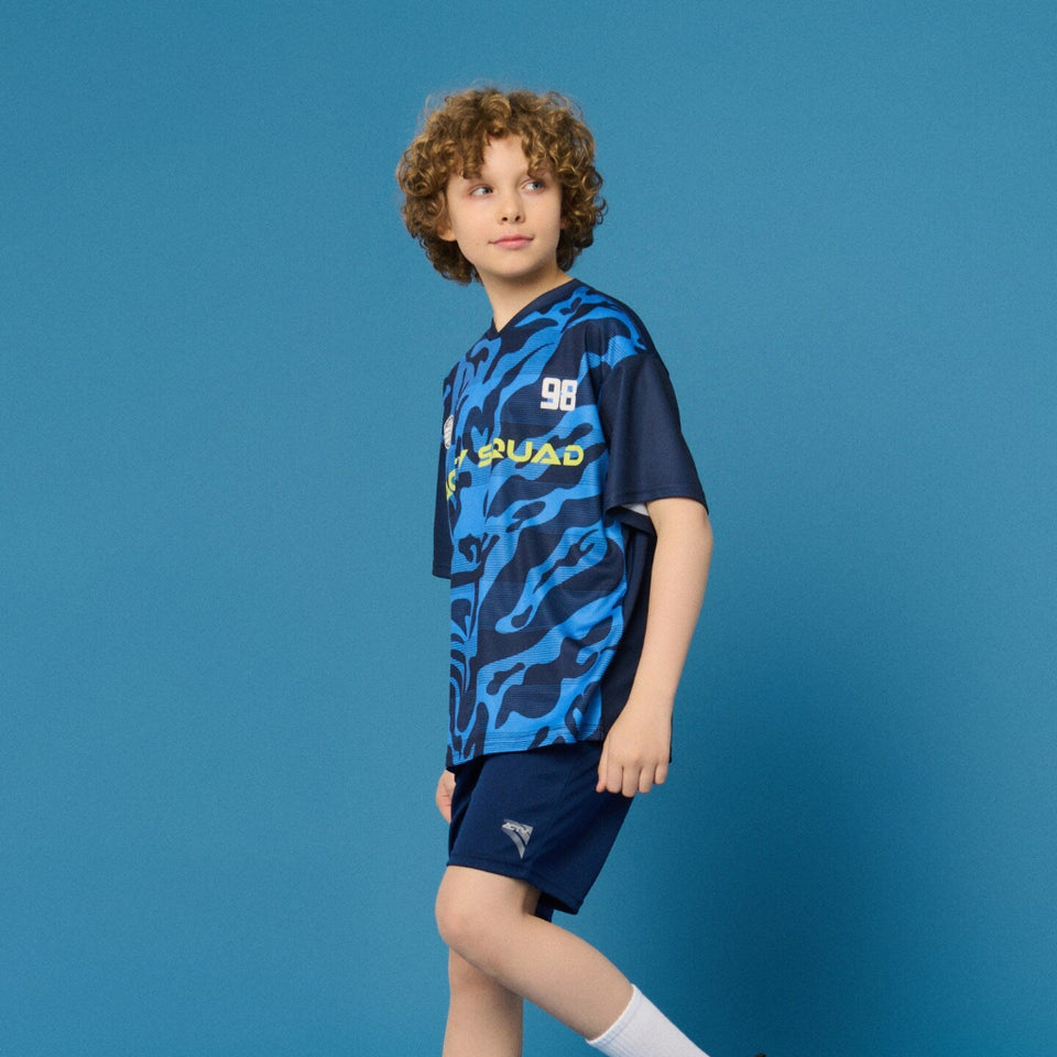 A child model wearing a blue and navy soccer jersey with a tiger-stripe pattern, navy shorts, white socks, and black sneakers, standing against a solid blue studio background.