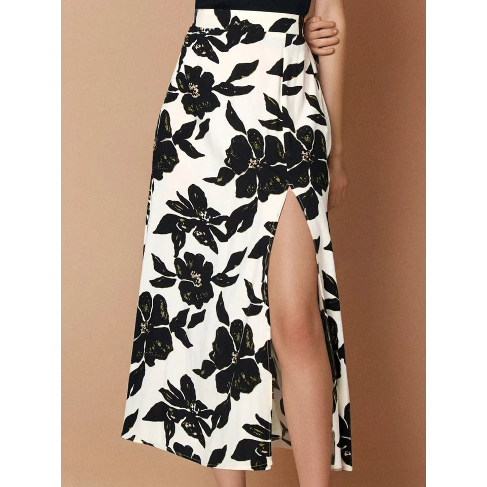 A high-waisted midi skirt with a black and white floral print and a thigh-high side slit, shown on a model against a solid tan background.