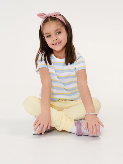 Young girl sitting on the floor wearing a striped shirt and yellow pants against a white background