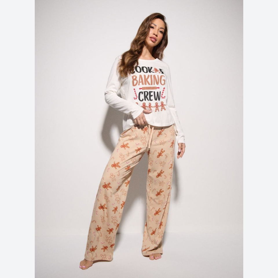 A woman modeling a two-piece pajama set consisting of a white long-sleeve crewneck shirt with a 'BOOK BAKING CREW' graphic and beige wide-leg pants with an orange gingerbread pattern, set against a plain light gray background.
