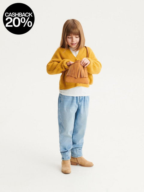 Child wearing a mustard yellow sweater and light blue jeans, holding a brown bag on a white background