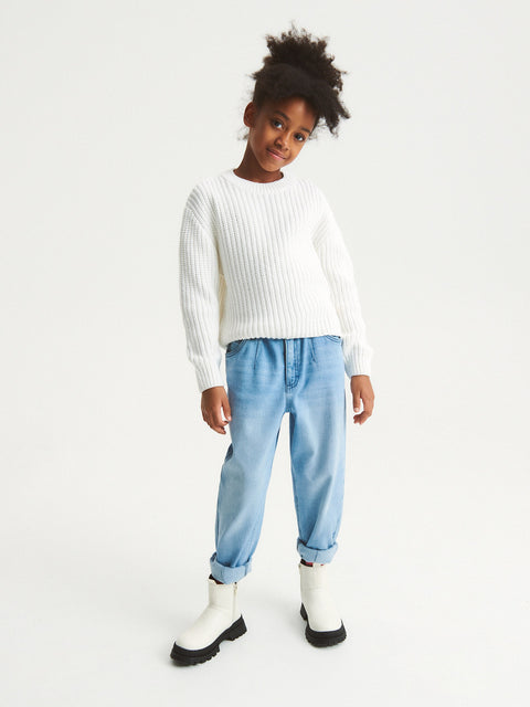 Child wearing a white sweater, light blue jeans, and black boots on a white background