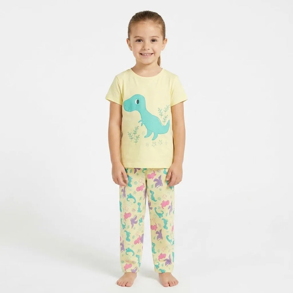 A young girl stands barefoot against a plain white studio background wearing a pale yellow pajama set with a large blue dinosaur graphic on the shirt and multicolored dinosaur prints on the pants.