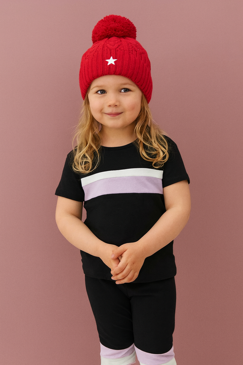 Child wearing a red knit hat with a star and a black outfit with a purple stripe on a brown background.