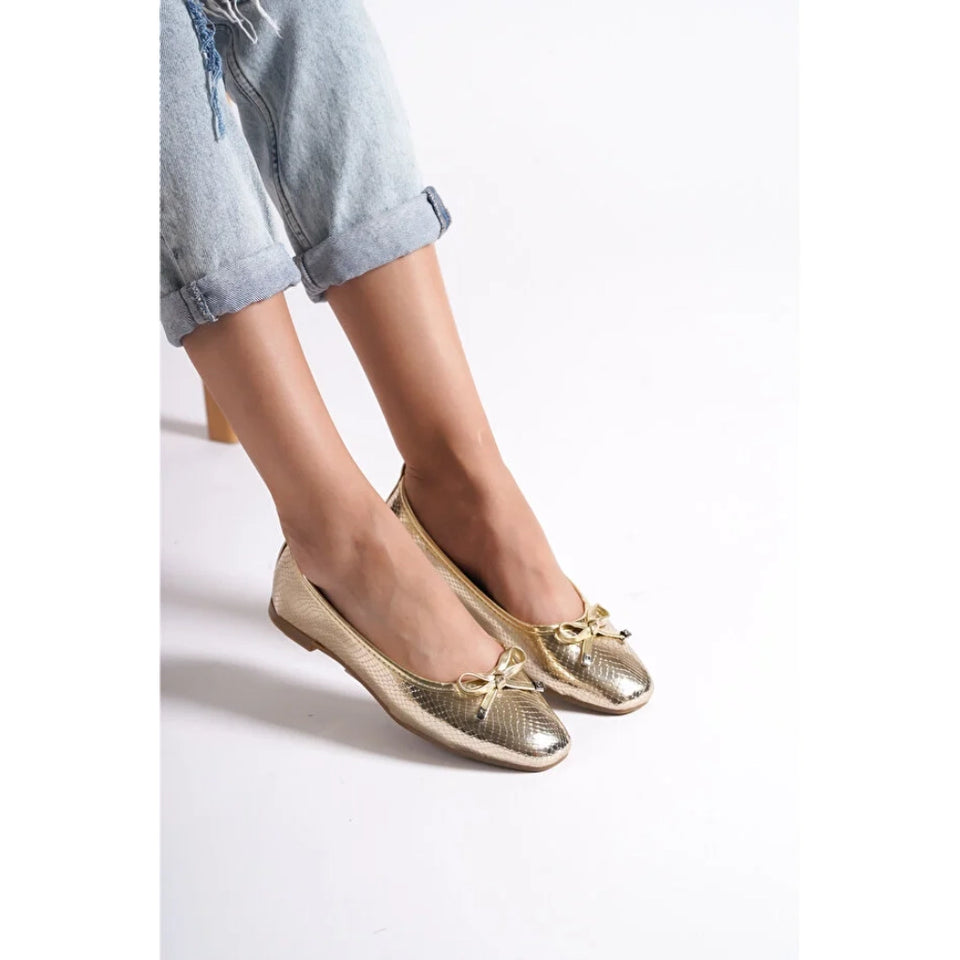 A person wearing metallic gold flat shoes with bow details and light blue rolled-up jeans, shown from the knees down on a plain white studio background.