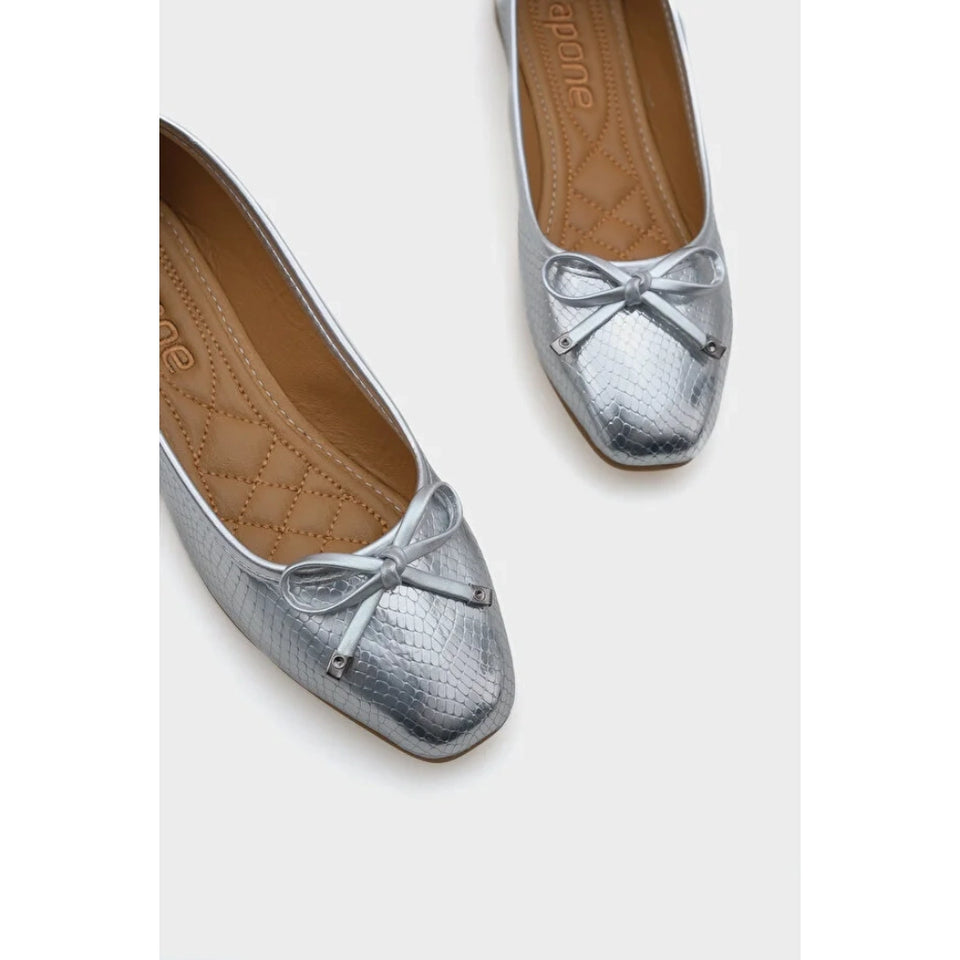 A pair of metallic silver ballet flats with a textured python-like finish and a small matching bow on the toe, shown on a light gray background.