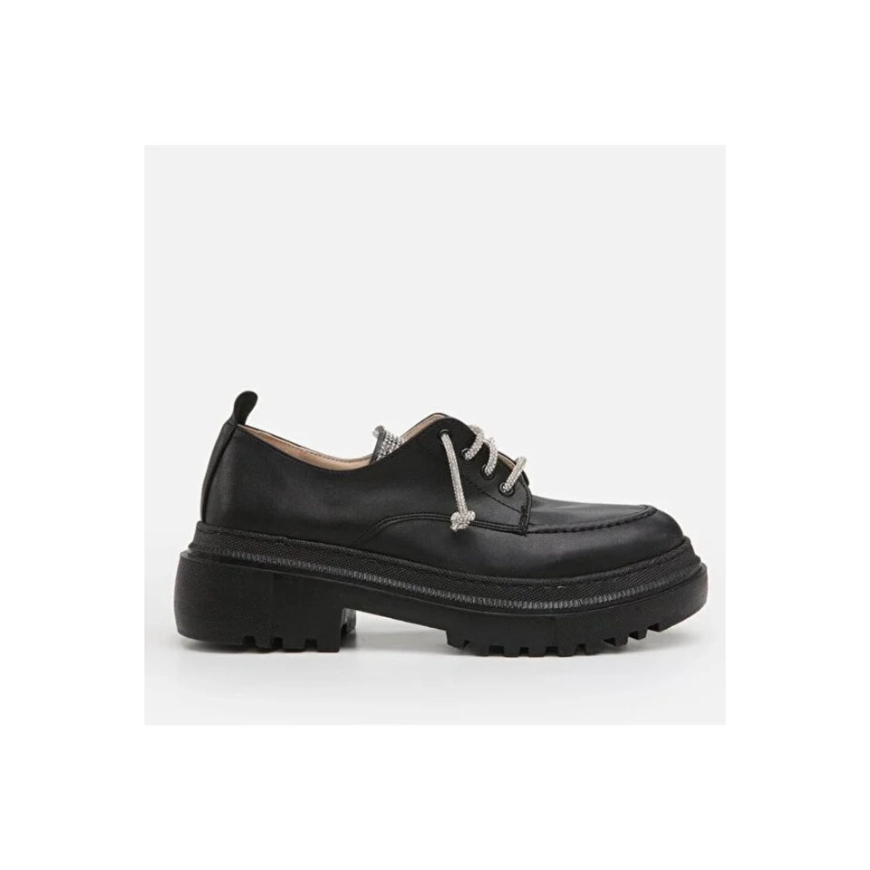 A single black leather oxford-style shoe with a chunky lug sole and silver-toned decorative laces, shown in profile against a light gray background.