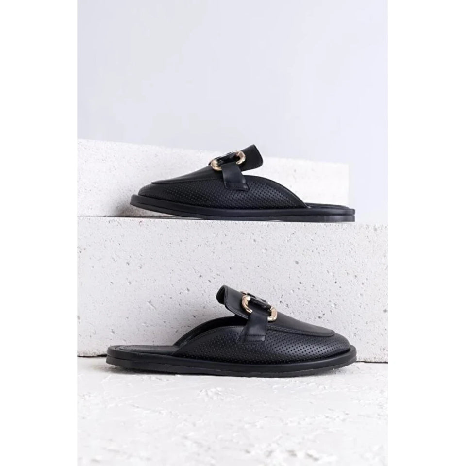 A pair of black leather slide sandals with a textured upper and a gold-tone buckle detail, displayed on white concrete steps against a light gray background.