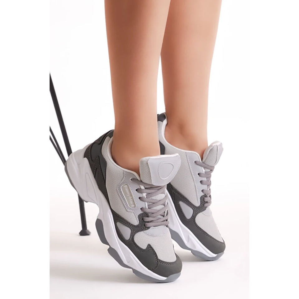 A pair of chunky low-top sneakers in gray, white, and black, shown on a model's feet against a clean white background.