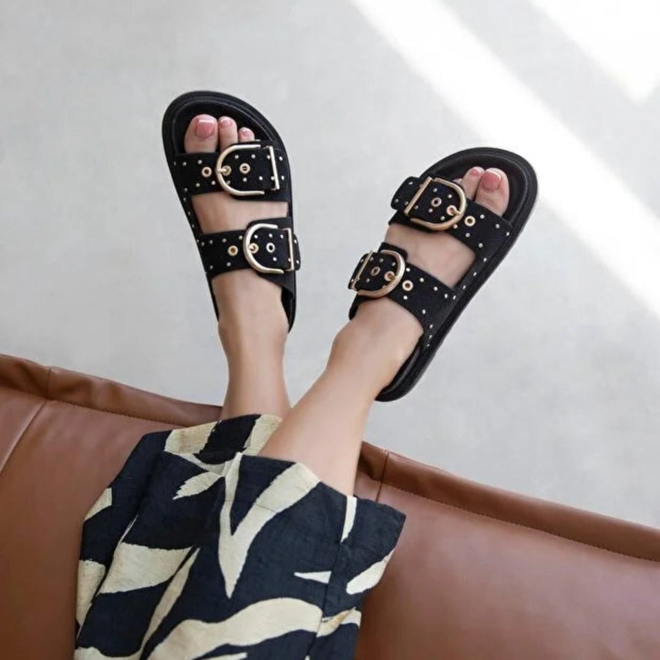 A person wearing black double-strap slide sandals with large gold-tone buckles and gold eyelet details, seated on a brown leather couch. The person is also wearing a navy and cream patterned skirt.