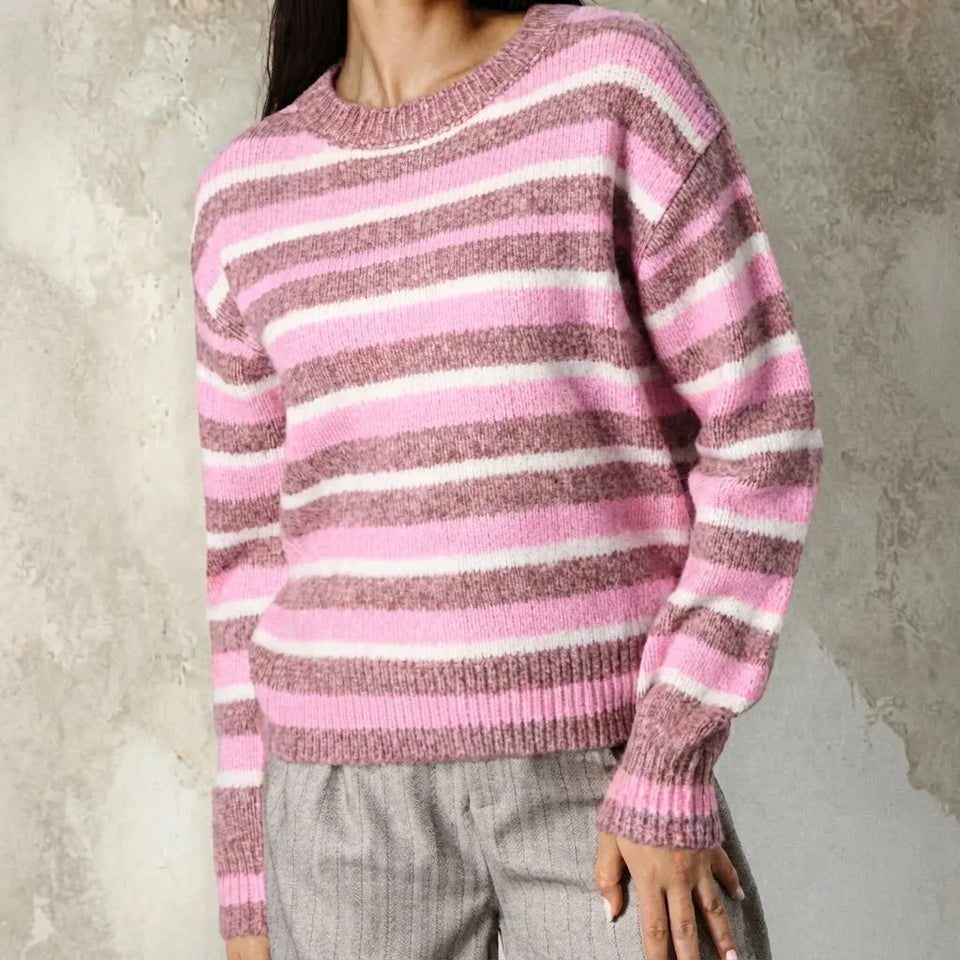 A person wearing a pink, white, and mauve striped knit sweater with gray pants, standing against a textured gray wall.