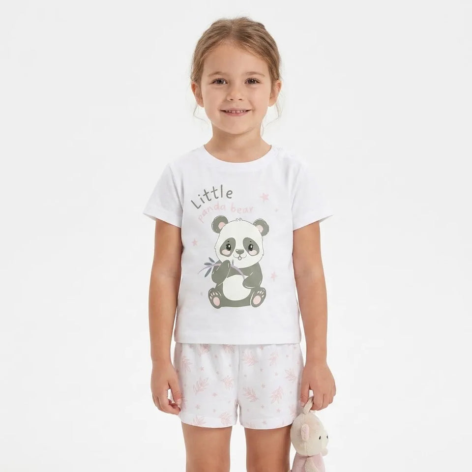 A young girl stands against a light gray studio background wearing a white short-sleeve pajama top with a panda graphic and the words 'Little panda bear,' matching white shorts with pale pink floral prints, and holding a small plush toy.