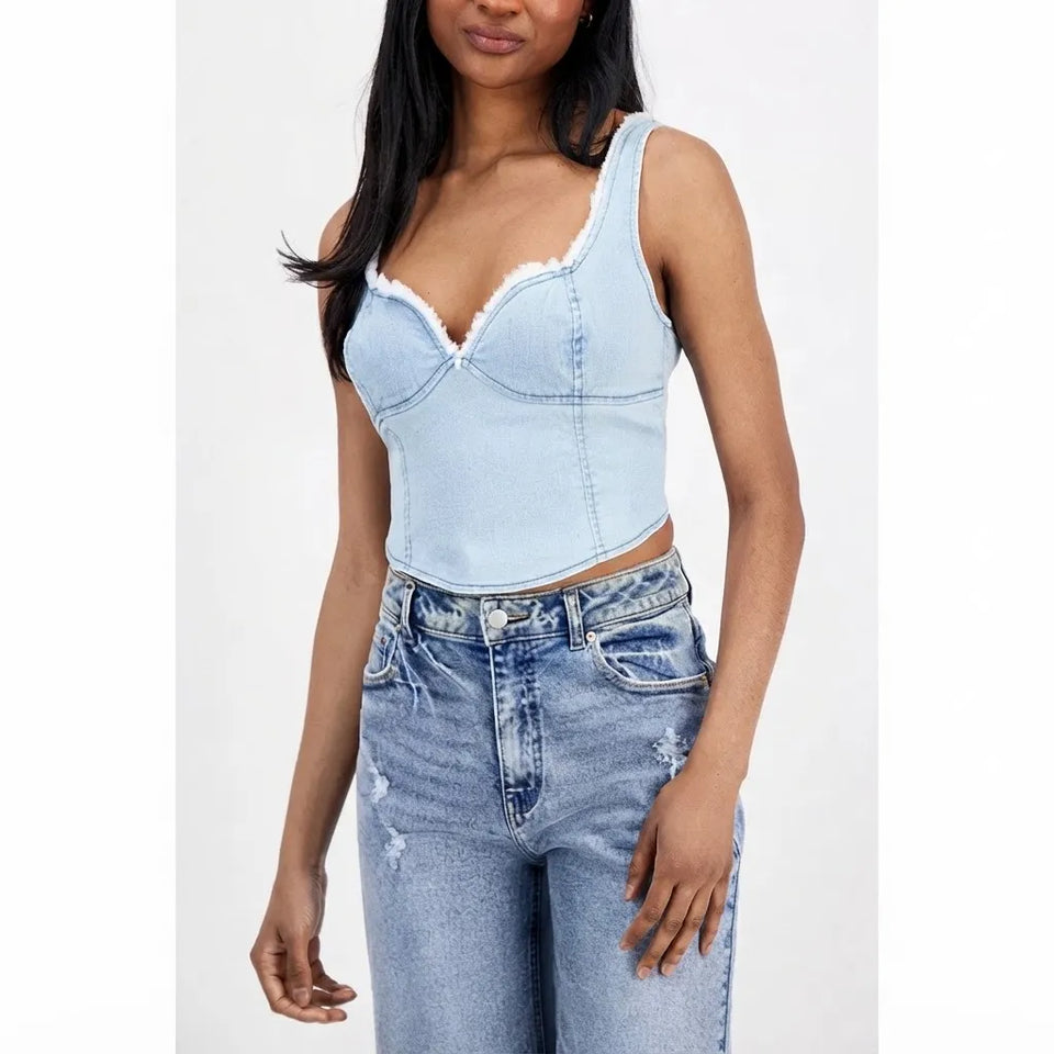 A woman wearing a light blue denim corset-style camisole top with lace-up front and ruffled trim, paired with light-wash distressed jeans, against a plain light gray background.