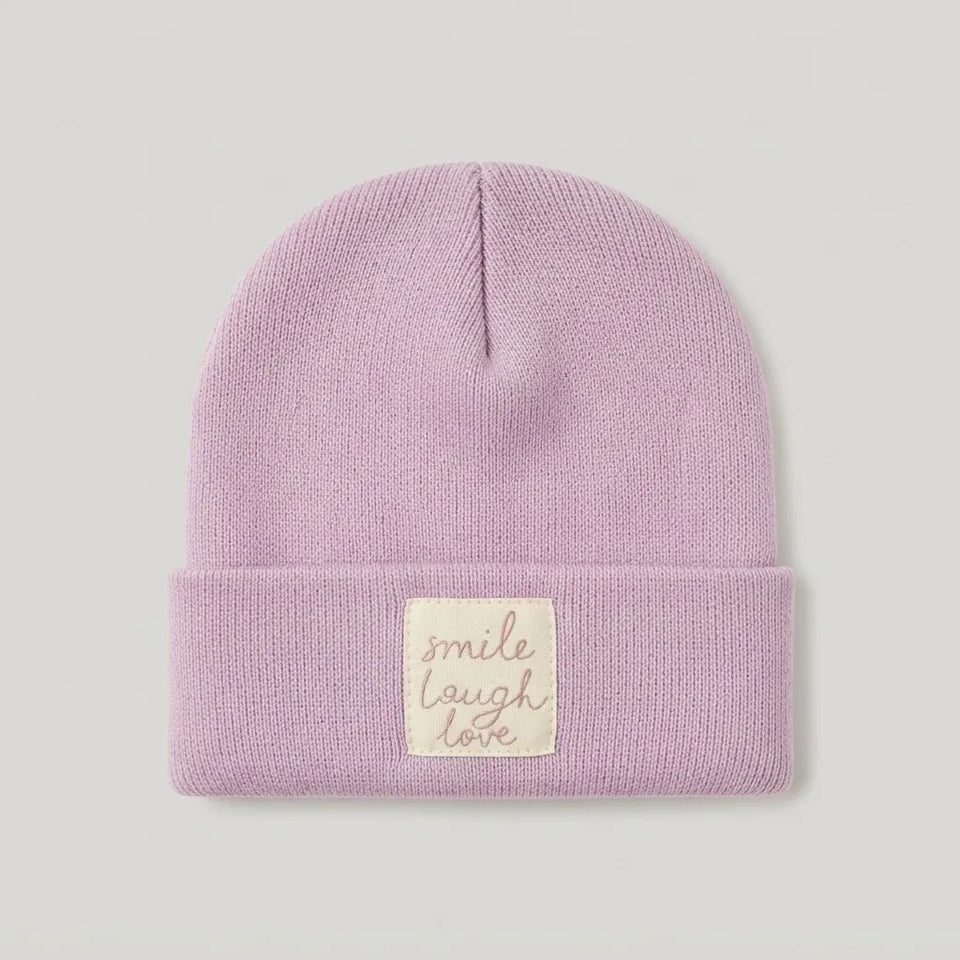 A light purple knit beanie hat with a folded cuff, featuring a cream-colored square patch with the embroidered words 'smile laugh love' in a cursive font, set against a neutral light gray background.