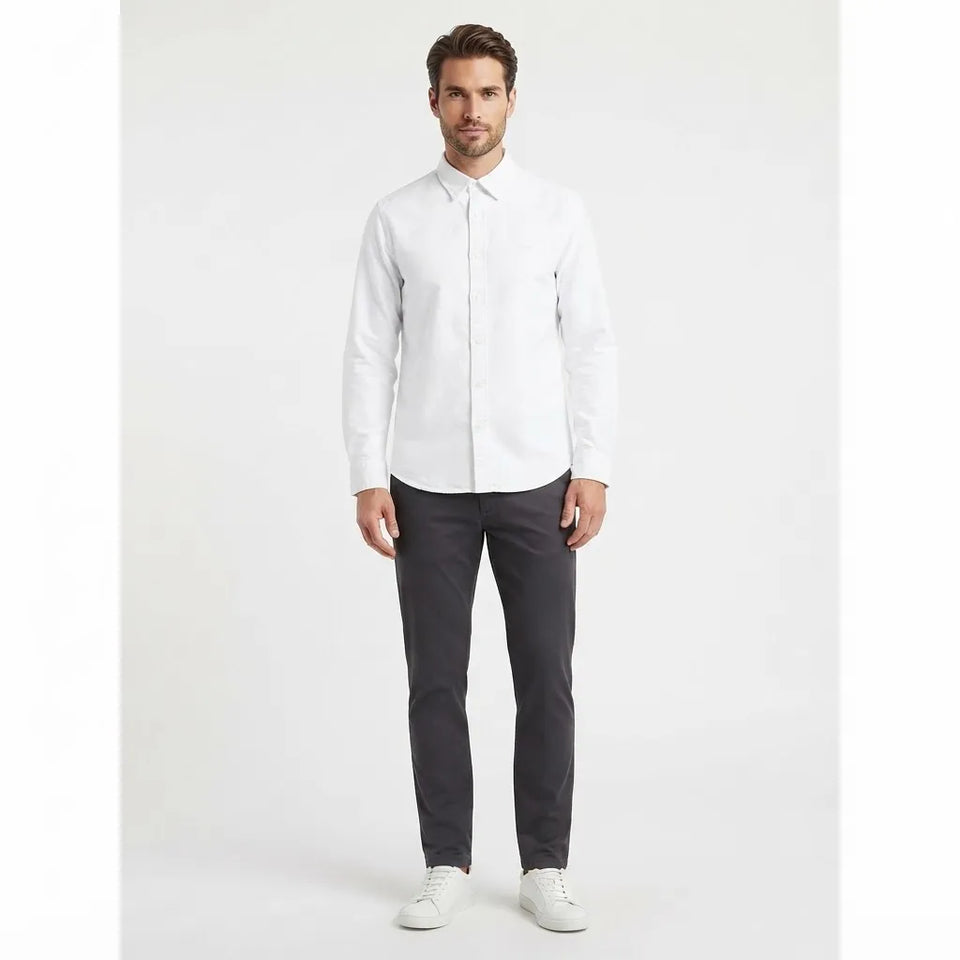 A man modeling a white long-sleeve button-down shirt paired with dark gray trousers and white sneakers, standing against a light gray studio background.
