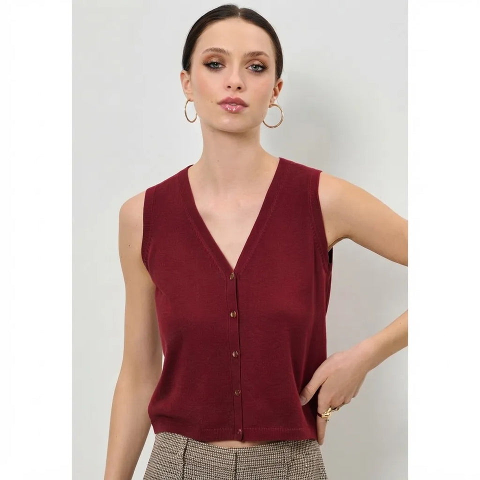 A woman modeling a sleeveless V-neck knit top in deep burgundy with a button-front closure, paired with patterned trousers, against a light gray studio background.