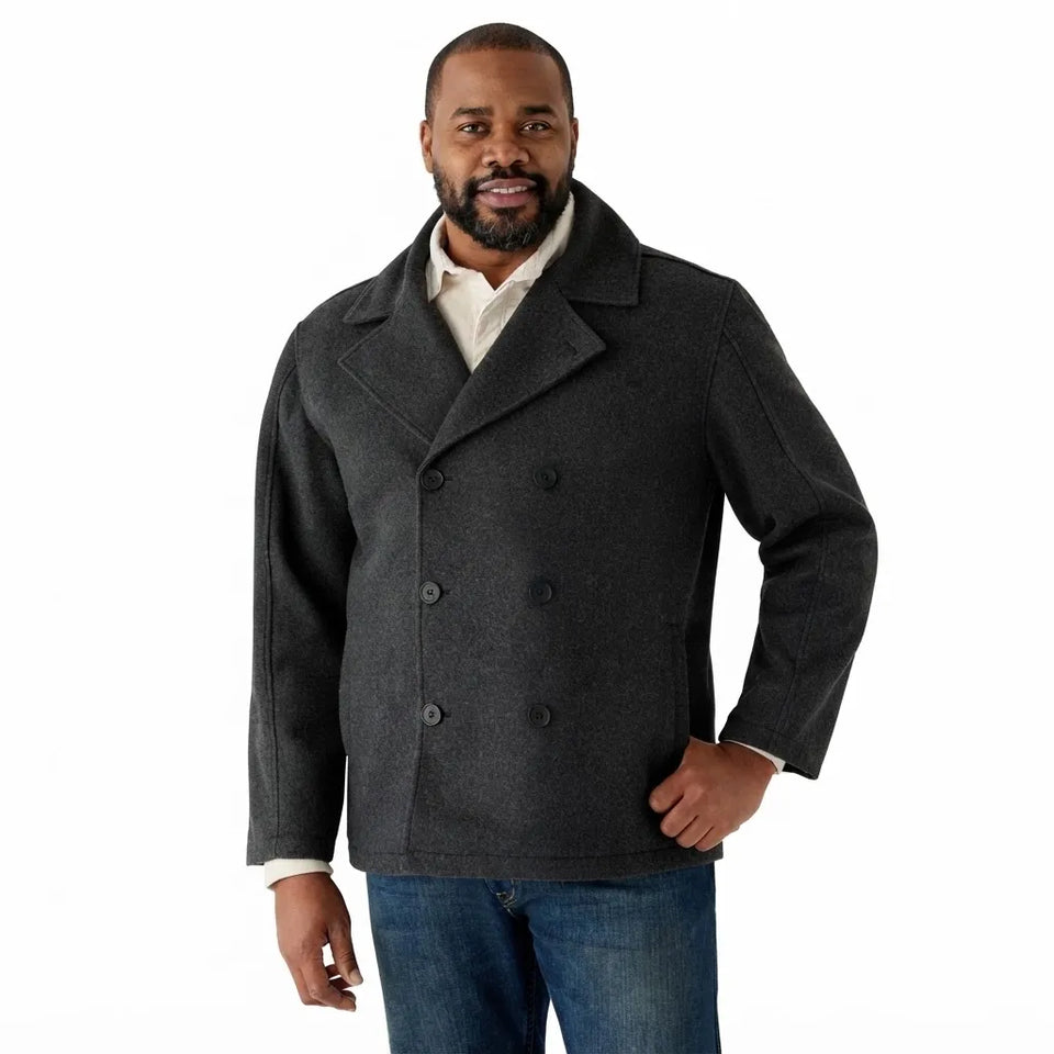 A man wearing a dark charcoal gray double-breasted peacoat over a cream shirt and blue jeans, photographed against a solid white background.