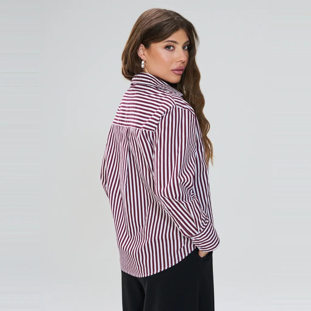 A woman wearing a long-sleeve button-up shirt with vertical burgundy and white stripes, paired with black pants, photographed against a light gray studio background.
