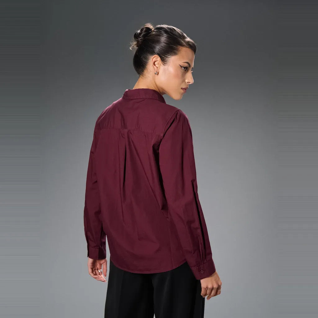 A woman shown from the back wearing a long-sleeve high-neck blouse in deep burgundy with black pants, photographed against a dark gray studio background.