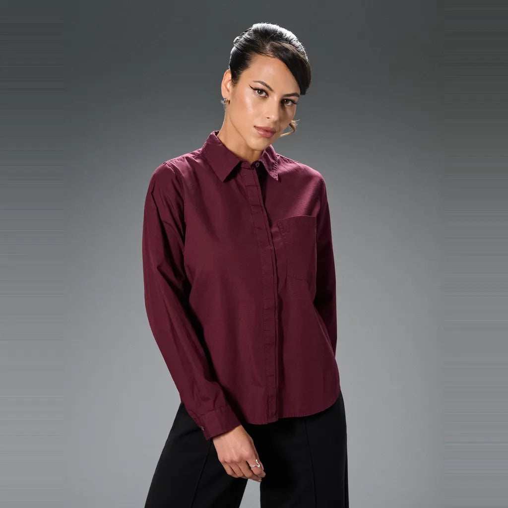 A woman modeling a long-sleeve button-up shirt in deep burgundy with a chest pocket, paired with black pants, against a dark gray studio background.