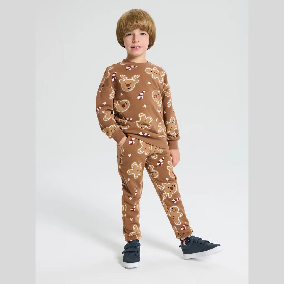 A young boy modeling a matching brown pajama set with a gingerbread-themed print, consisting of a long-sleeve sweatshirt and jogger pants, paired with dark blue sneakers against a light gray studio background.