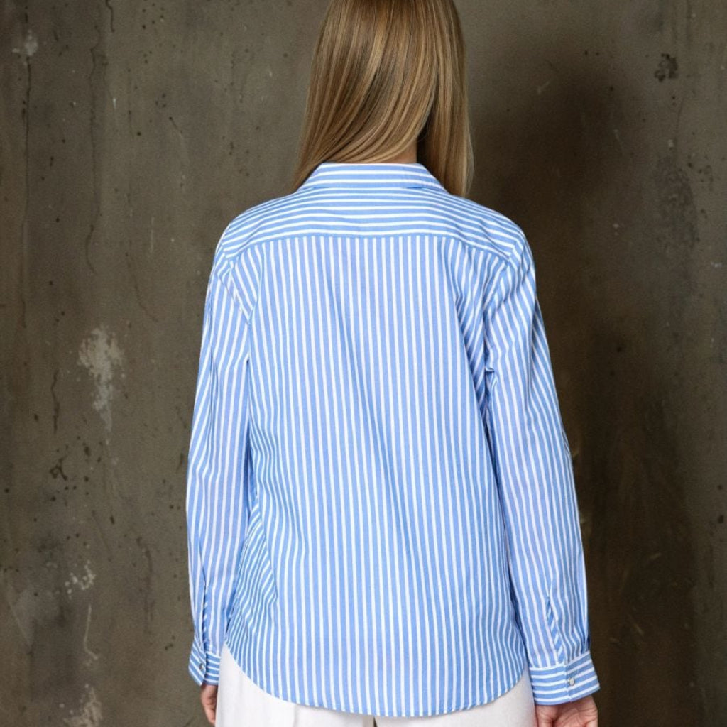 A woman viewed from behind wearing a light blue and white vertically striped button-down shirt and white trousers, standing against a textured concrete wall.