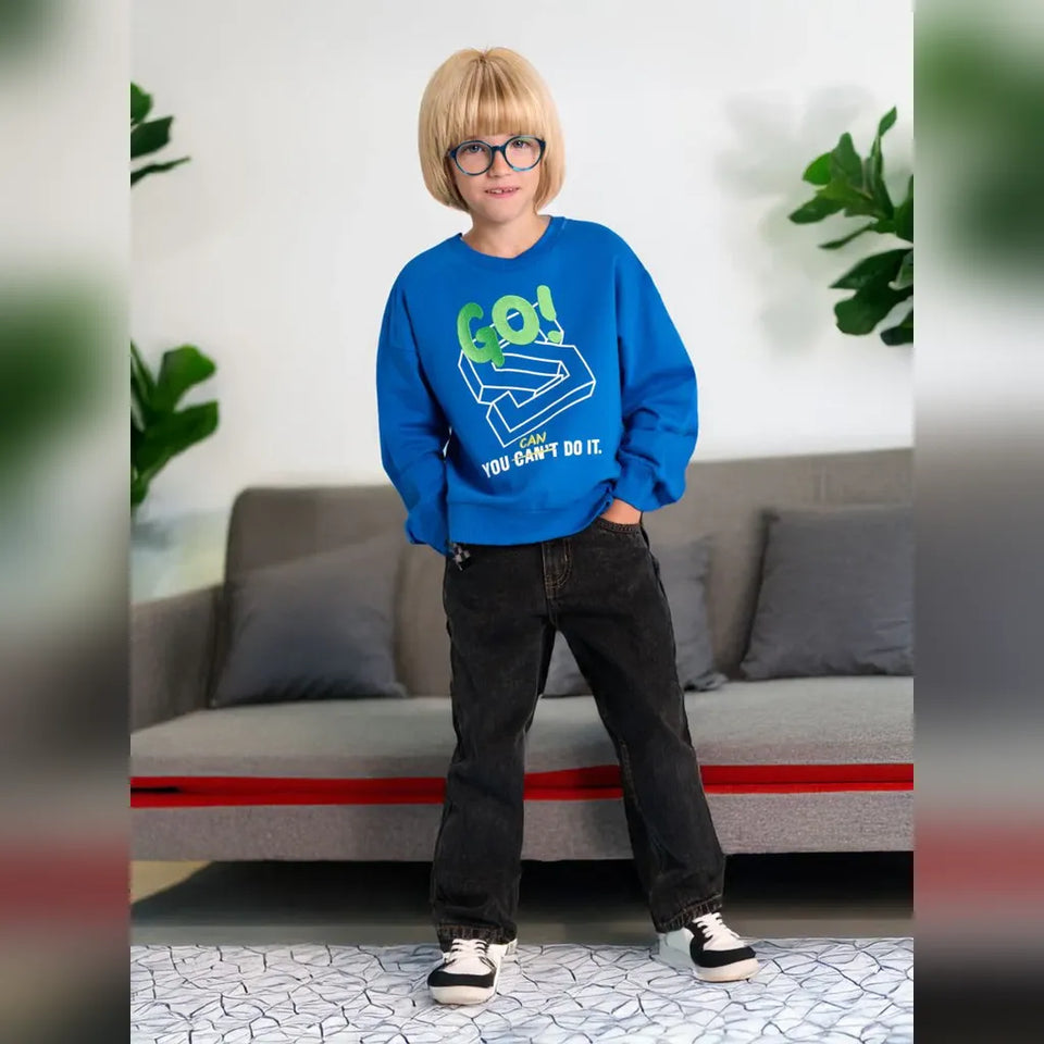 A child wearing a bright blue sweatshirt with a green and white graphic that reads 'Go! YOU CAN'T DO IT.' paired with dark jeans and white sneakers, standing in front of a gray sofa in a living room setting.