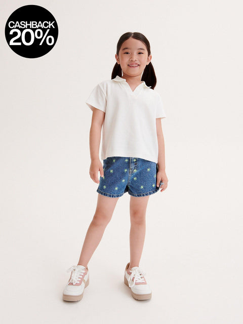 Young girl wearing a white shirt and denim shorts with star patterns on a plain background