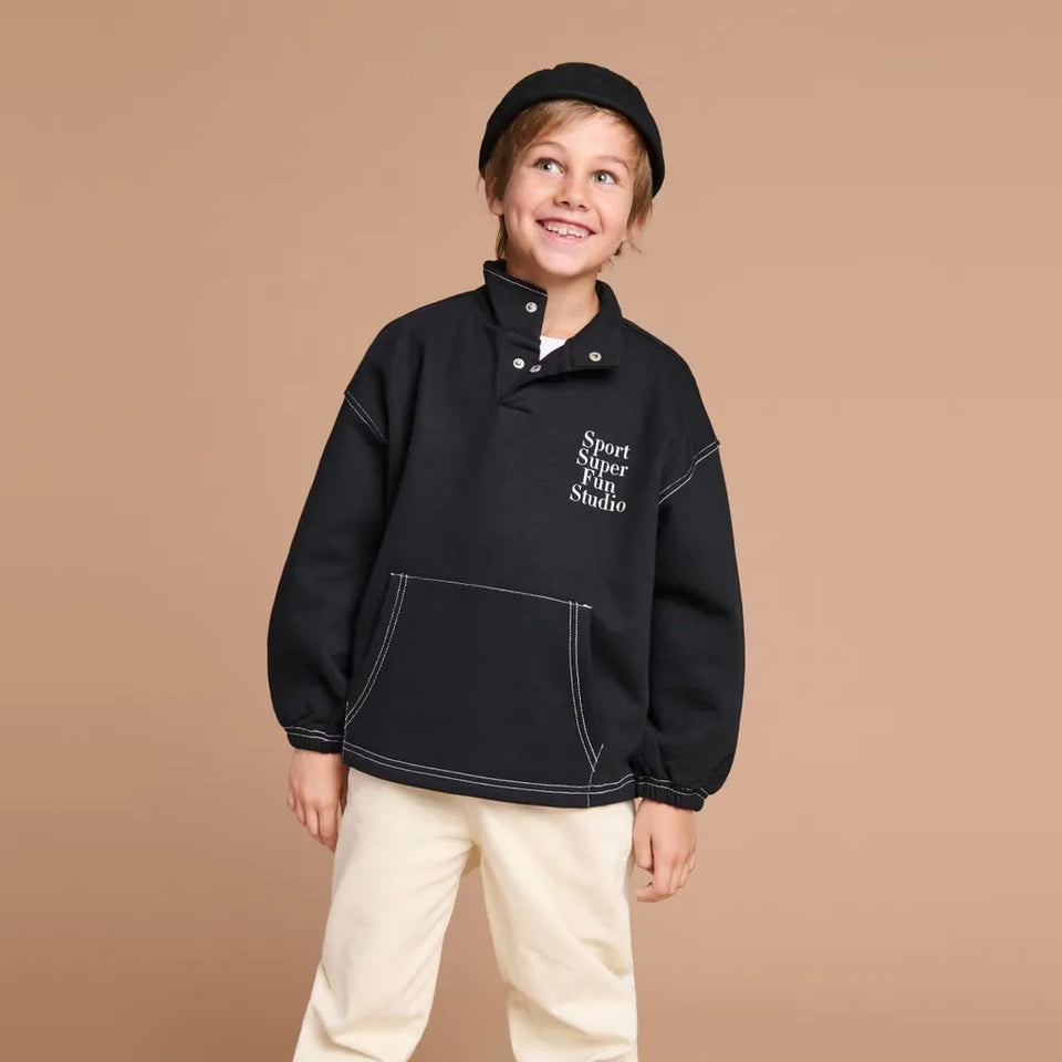 A young child wearing a black half-placket pullover sweatshirt with white contrast stitching and 'Sport Star Play Studio' text on the chest, paired with cream pants and a black beret, against a solid tan studio background.
