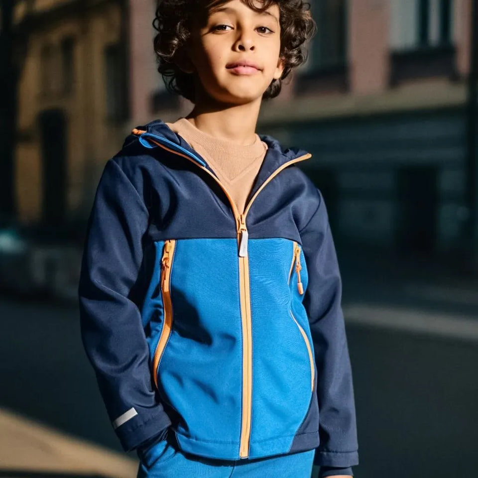 A child wearing a blue and navy zip-up hooded jacket with orange zipper accents, photographed outdoors in warm sunlight against a blurred street background.