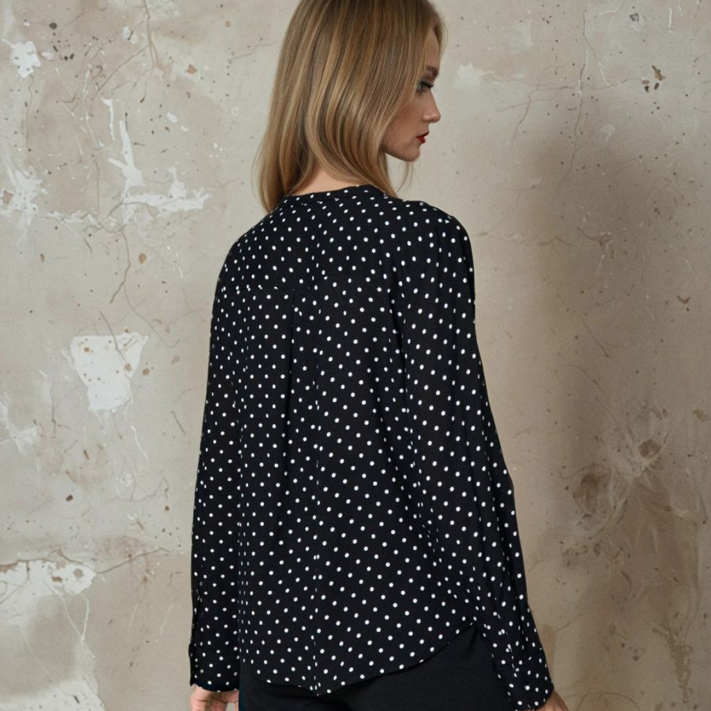 A woman shown from the back wearing a black long-sleeve blouse with a small white polka dot pattern, paired with black pants, standing against a textured beige plaster wall.