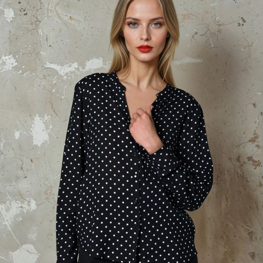 A woman modeling a black long-sleeve blouse with a white polka dot pattern, paired with black pants, standing against a textured concrete wall.
