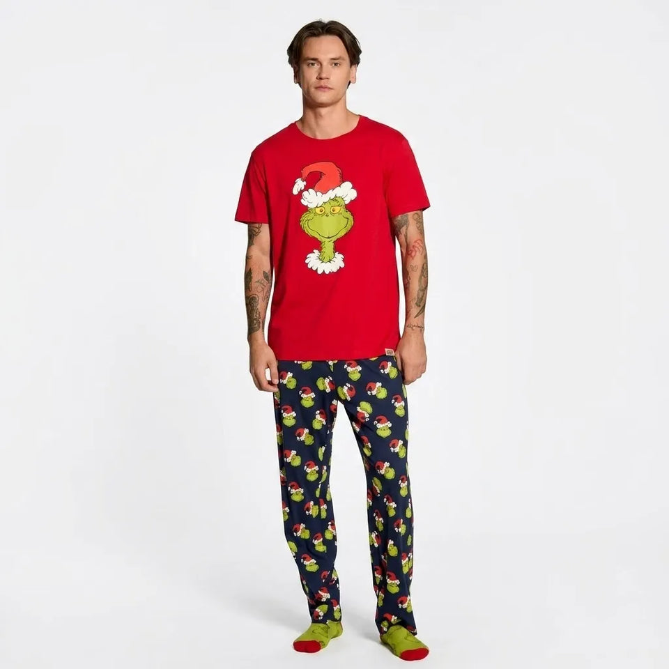 A man modeling a red short-sleeve t-shirt and dark navy patterned pajama pants with a Grinch-themed holiday print, standing against a plain white studio background.