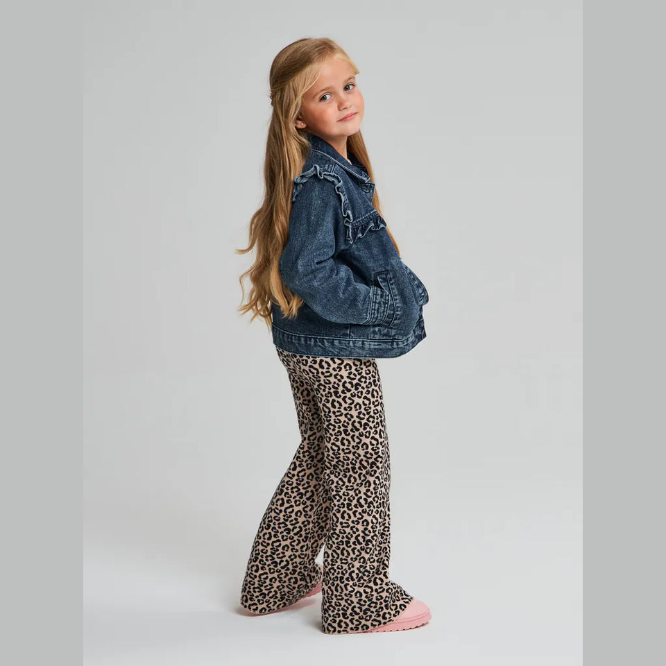 A young girl with long blonde hair stands in profile wearing a blue denim jacket, leopard-print pants, and pink slippers against a light gray studio background.